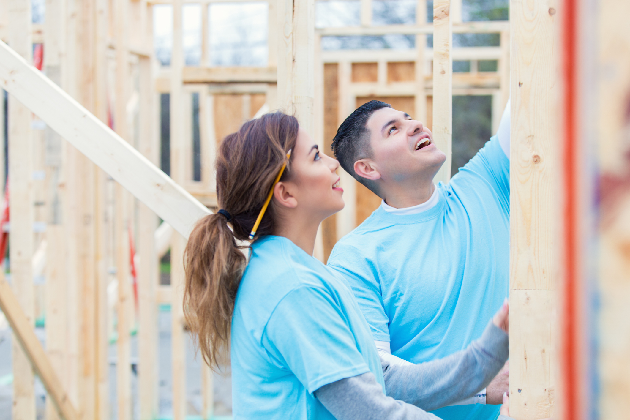 Couple volunteer with Habitat for Humanity. The man shows the woman something on a piece of plywood. They are wearing blue volunteer t-shirts. The woman has a pencil behind her ear. They are working on the frame for the house.